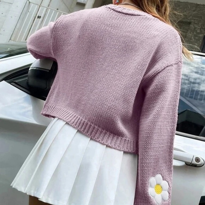 Paarse gebreide trui met bloemapplicatie op mouw, gedragen door vrouw naast witte auto. Modieuze dameskleding, trendy herfstoutfit.