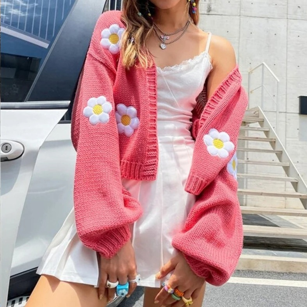 Vrouw in witte jurk met roze gebreid vest met bloemenpatroon, poseert buiten bij auto. Trendy damesmode, zomeroutfit, casual stijl.
