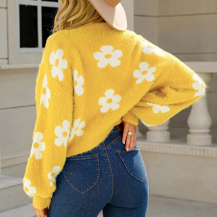 Vrouw in gele trui met witte bloemen, draagt jeans en hoed, buiten bij gebouw. Trendy herfstmode, casual outfit, stijlvolle dameskleding.