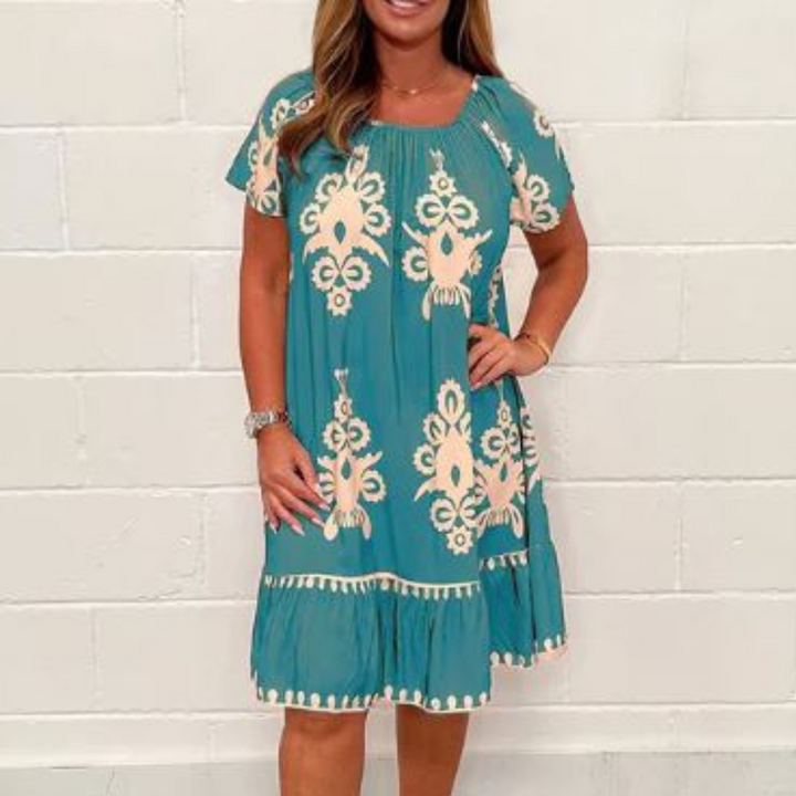 Vrouw draagt blauwe zomerjurk met witte patronen, korte mouwen en ronde hals. Perfect voor casual en stranduitjes. Comfortabele en stijlvolle damesmode.