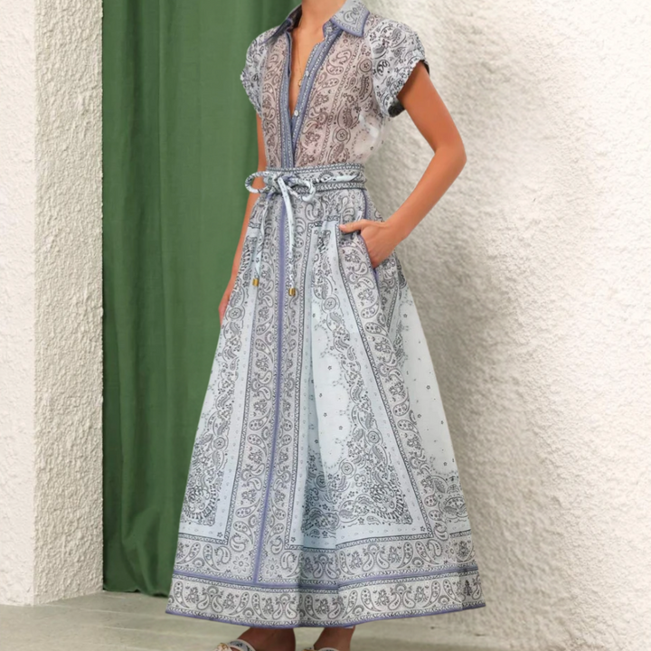 Vrouw in blauwe paisley maxi-jurk met korte mouwen en ceintuur, staand voor groene en witte achtergrond. Elegante zomerjurk mode.