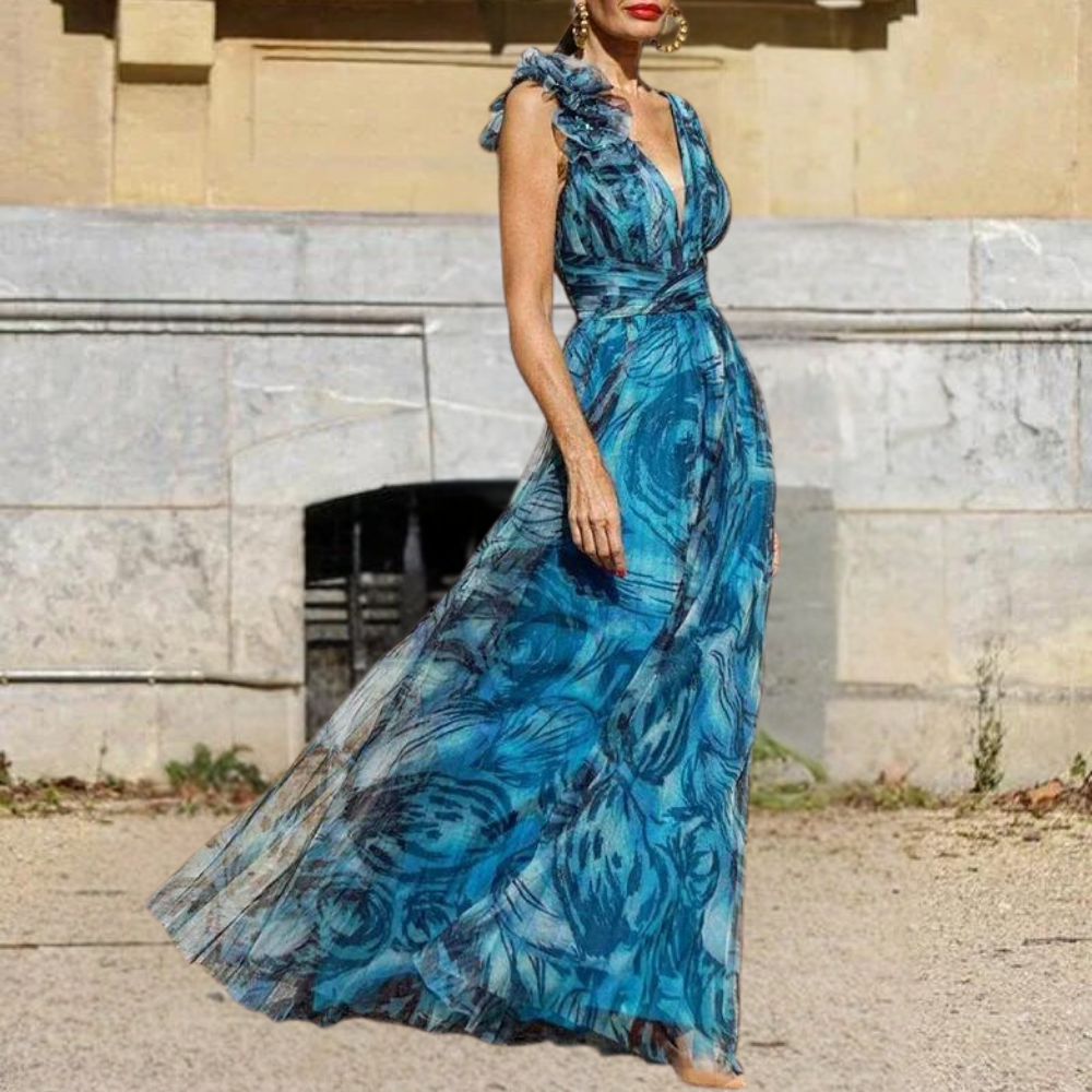 Vrouw in elegante blauwe bloemenjurk met ruches, staand voor historische stenen muur. Perfect voor zomerfeestjes en bruiloften.