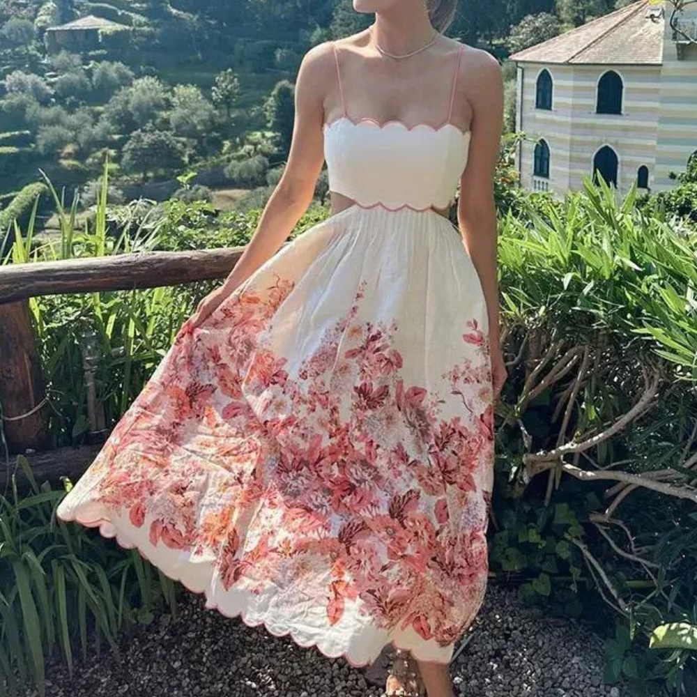 Vrouw in zomerse witte jurk met roze bloemenprint, spaghetti bandjes en golvende zoom, buiten in een groene tuin. Perfect voor zomermode.