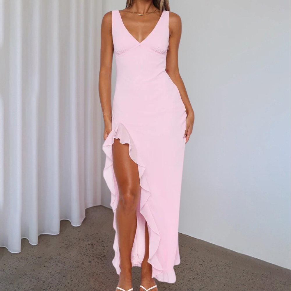Vrouw in elegante roze avondjurk met diepe V-hals en hoge split, staand voor witte gordijnen. Perfect voor galafeest of bruiloft.