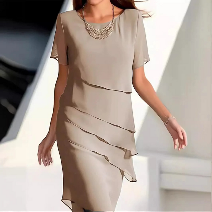 Vrouw in elegante, beige jurk met gelaagde details en korte mouwen, draagt een ketting. Perfect voor formele gelegenheden. Modieuze dameskleding.