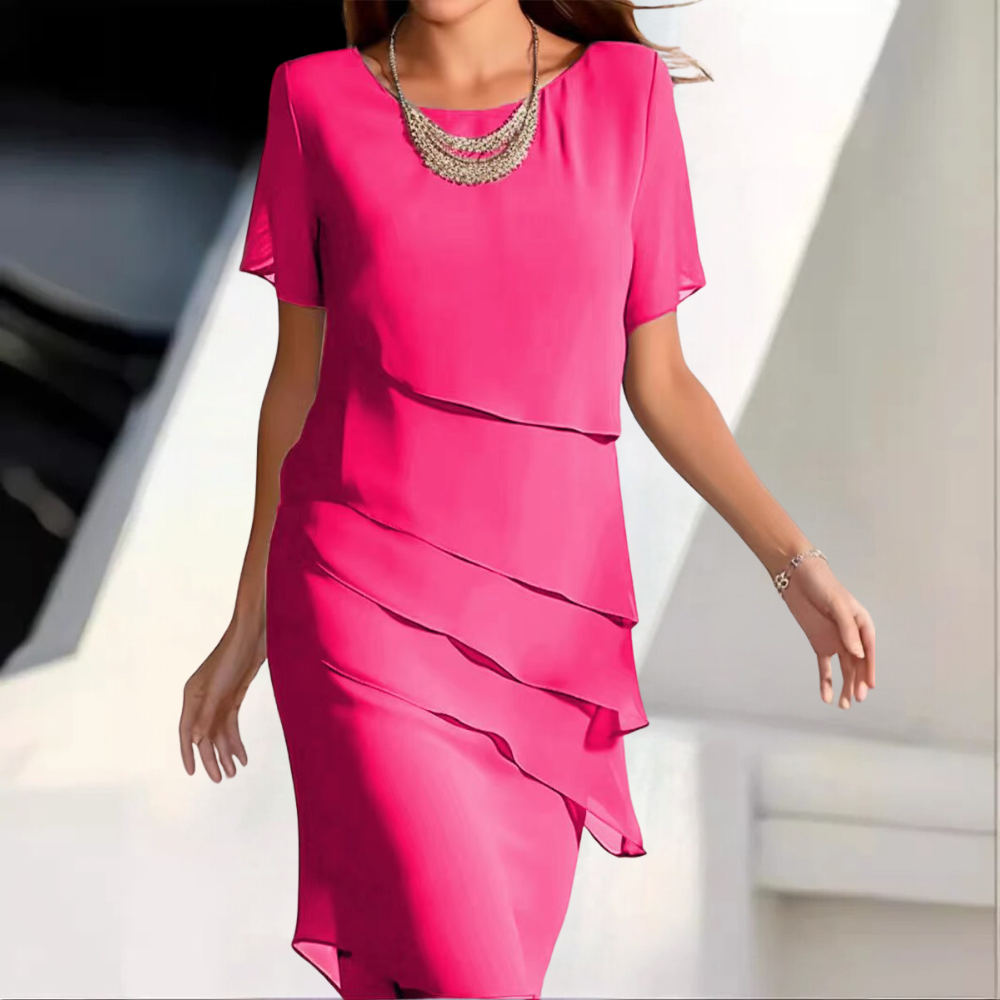 Vrouw in elegante roze jurk met gelaagde details, korte mouwen en opvallende ketting. Perfect voor feestelijke gelegenheden en zomermode.
