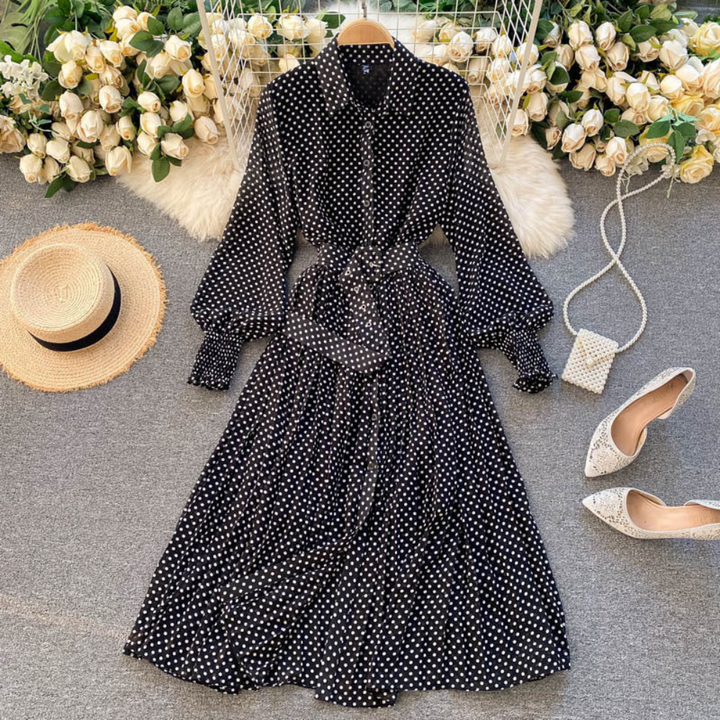 Zwarte polkadot jurk met lange mouwen en strikceintuur, omgeven door witte rozen, hoed, witte hakken en een kleine tas. Perfect voor elegante gelegenheden.