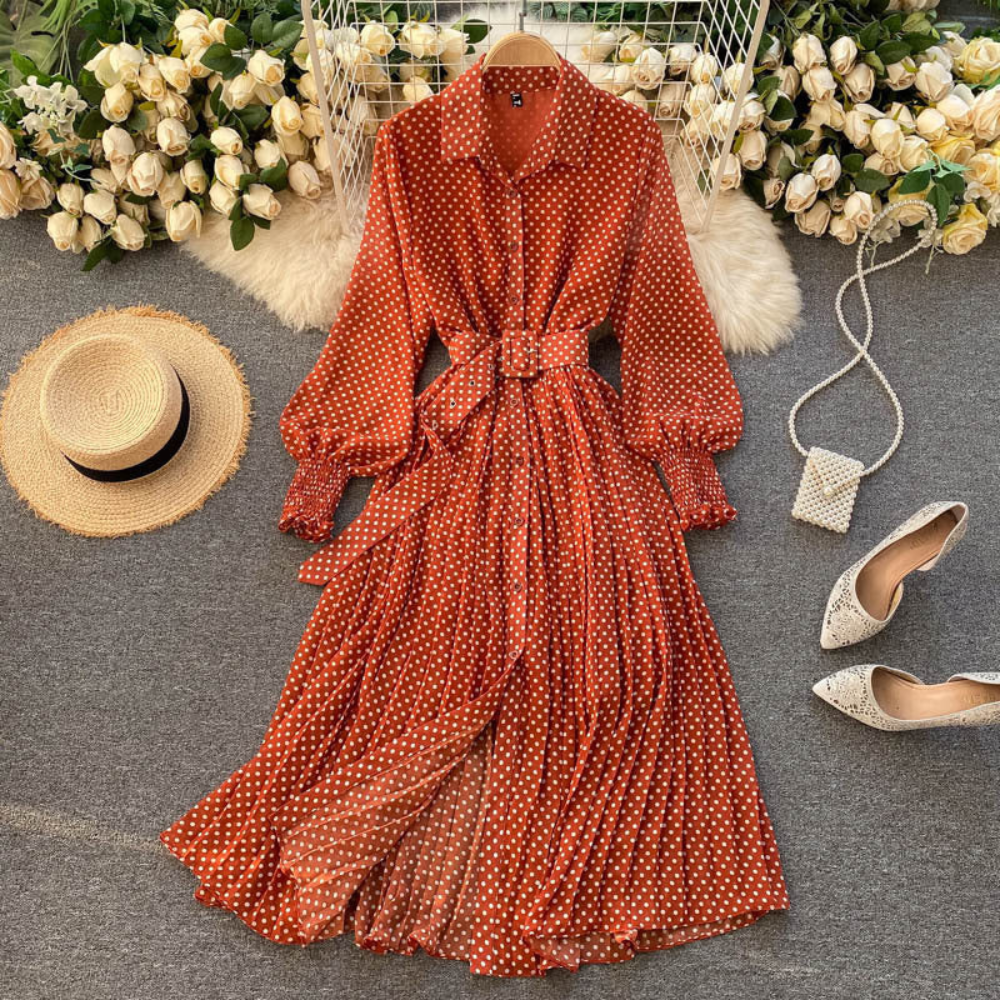 Oranje-rode polkadot maxi-jurk met lange mouwen en ceintuur, omgeven door witte rozen, strohoed, witte hakken en een kleine tas.