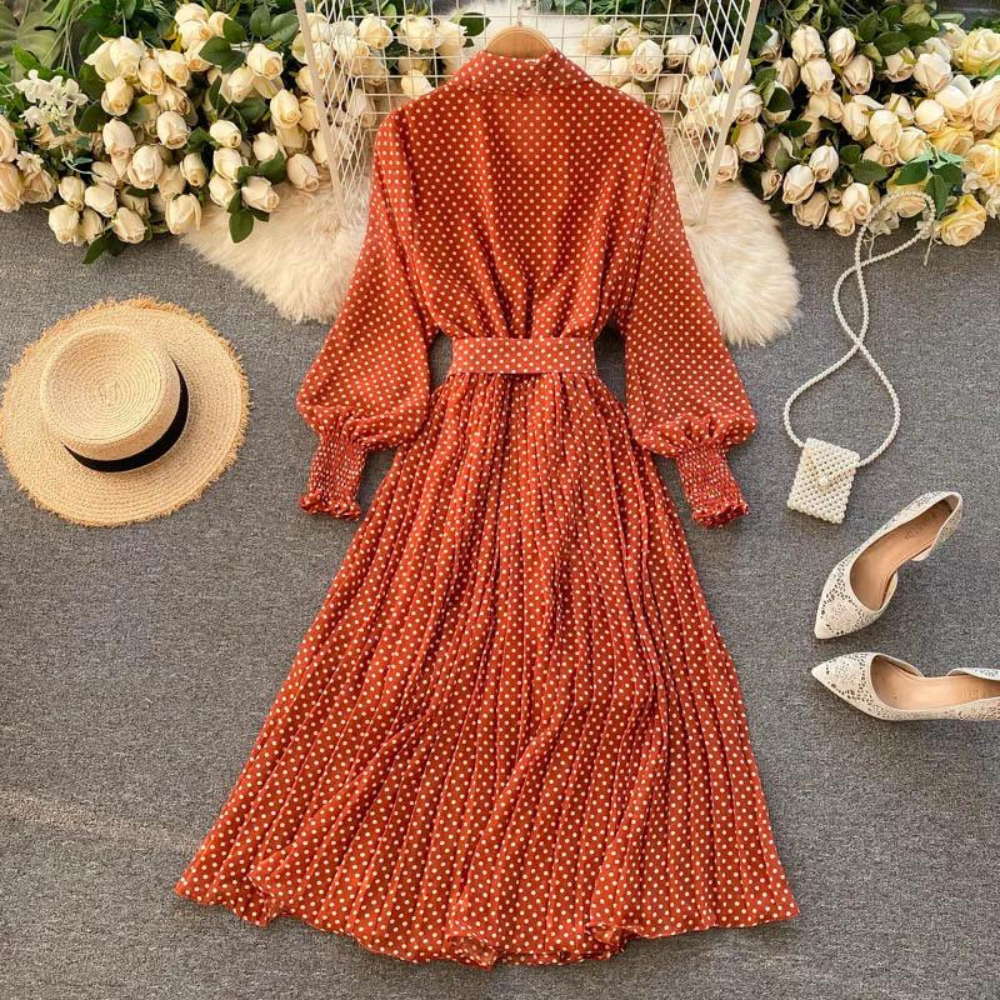 Oranje-rode polkadot jurk met lange mouwen en ceintuur, omgeven door witte rozen, strohoed, witte hakken en een kleine handtas op grijze achtergrond.