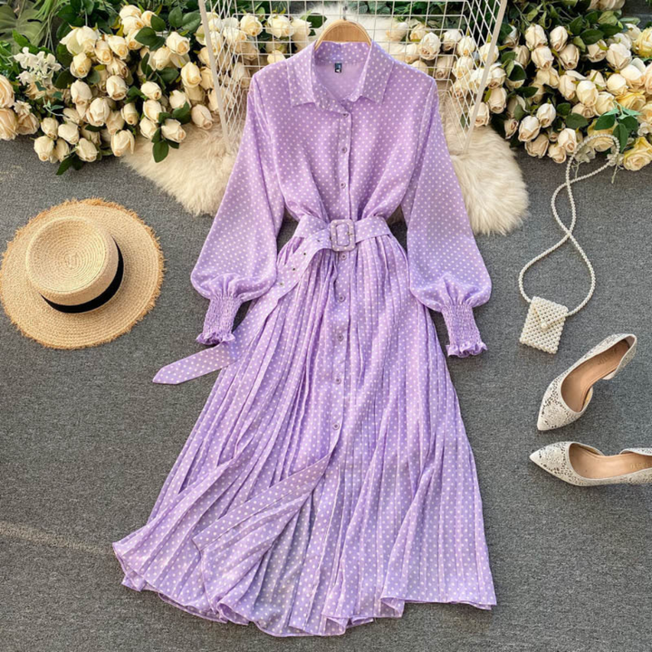 Lange paarse jurk met stippen en ceintuur, omgeven door witte rozen, hoed en schoenen. Perfect voor lente- of zomermode.