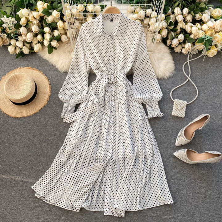 Witte polkadot maxi-jurk met lange mouwen, naast een strohoed, parelhandtas en puntige schoenen, omringd door witte rozen. Perfect voor zomermode.