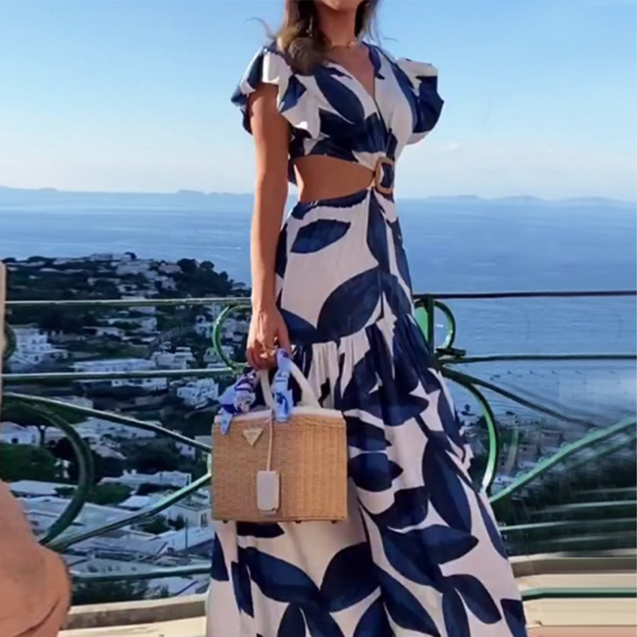 Vrouw in blauwe en witte maxi-jurk met bladmotief, draagt een rieten tas. Achtergrond: panoramisch uitzicht op zee en stad. Zomermode, strandkleding.