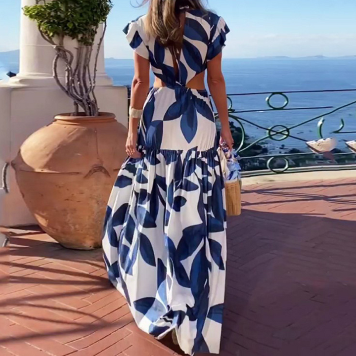 Vrouw in lange blauwe en witte zomerjurk met bladpatroon, staand op terras met zeezicht. Perfecte outfit voor strandvakantie of zomerse bruiloft.