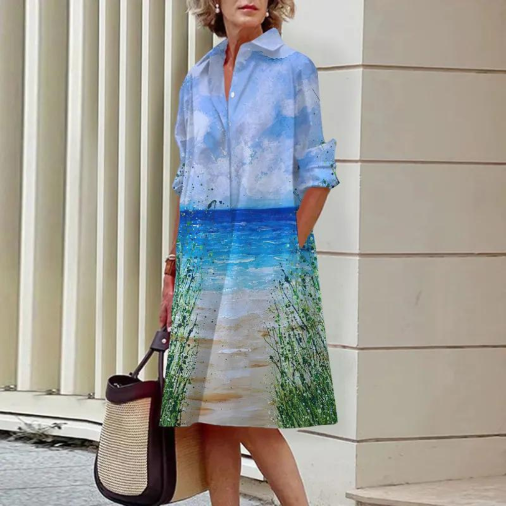 Vrouw in strandjurk met zee- en luchtprint, draagt een rieten tas. Perfect voor zomermode, casual strandkleding, stijlvolle damesoutfit.