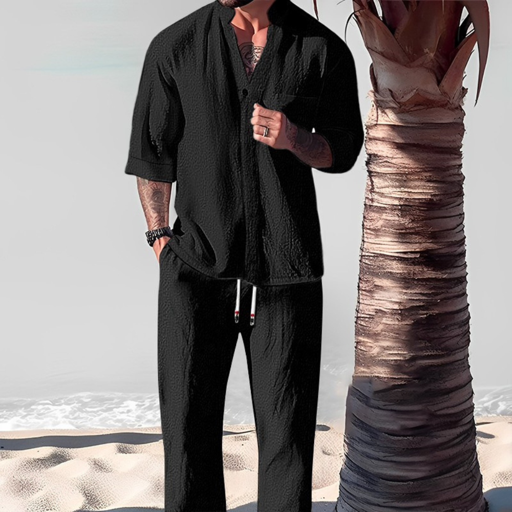 Man in zwarte linnen strandoutfit met korte mouwen en broek, naast palmboom op zandstrand. Perfect voor zomermode en strandvakanties.