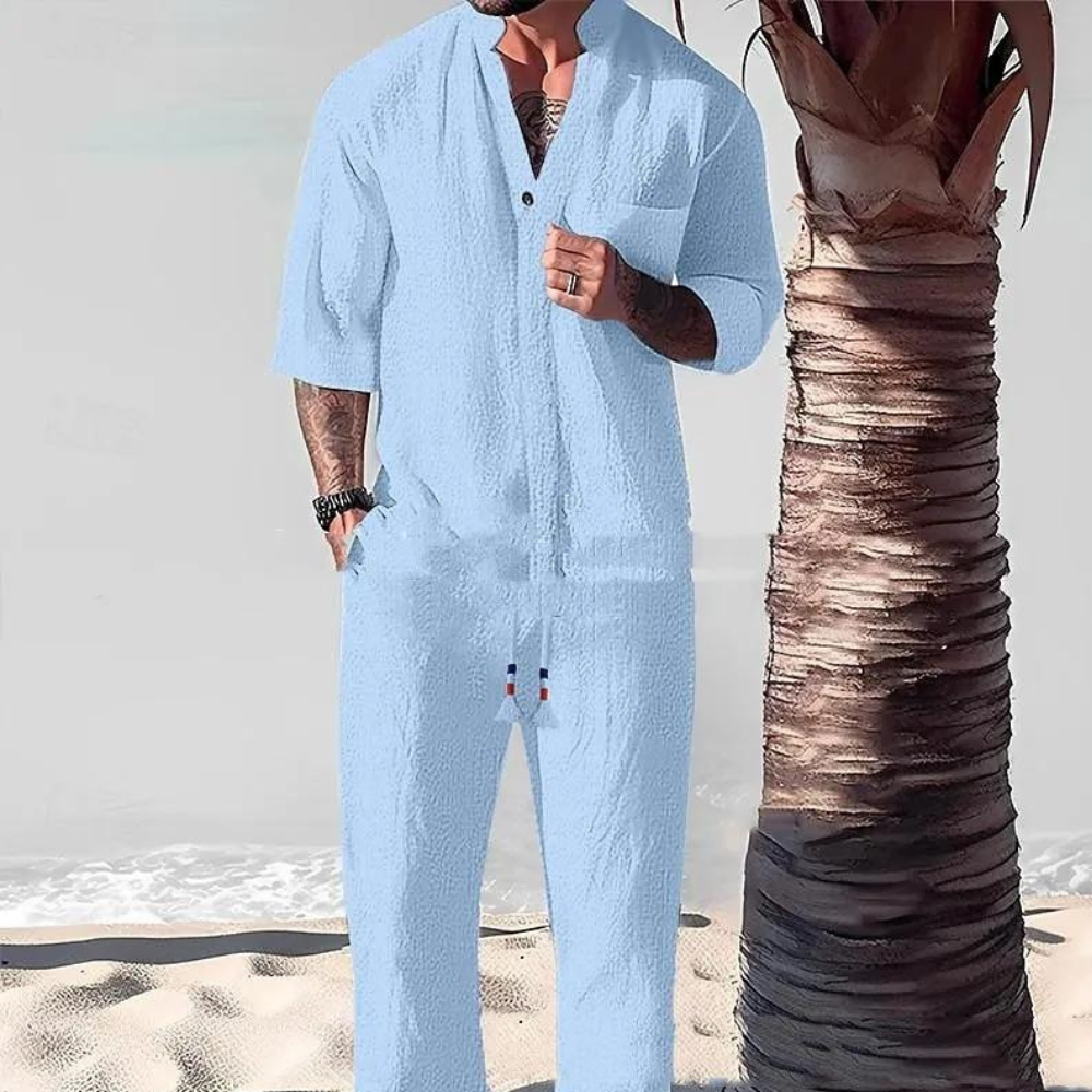 Man in lichtblauw linnen jumpsuit met korte mouwen, staand naast een palmboom op een zandstrand. Zomermode, casual strandkleding voor heren.