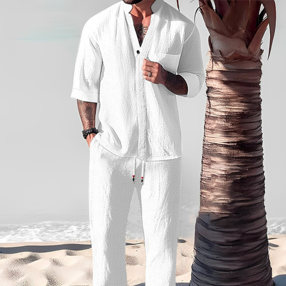 Man in witte linnen outfit op strand naast palmboom, zomermode, casual herenkleding, strandkleding, stijlvolle witte set, ontspannen sfeer.