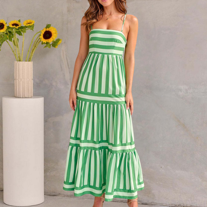 Vrouw in groene en witte gestreepte zomerjurk voor een grijze muur, naast een vaas met zonnebloemen. Perfecte outfit voor zomermode.