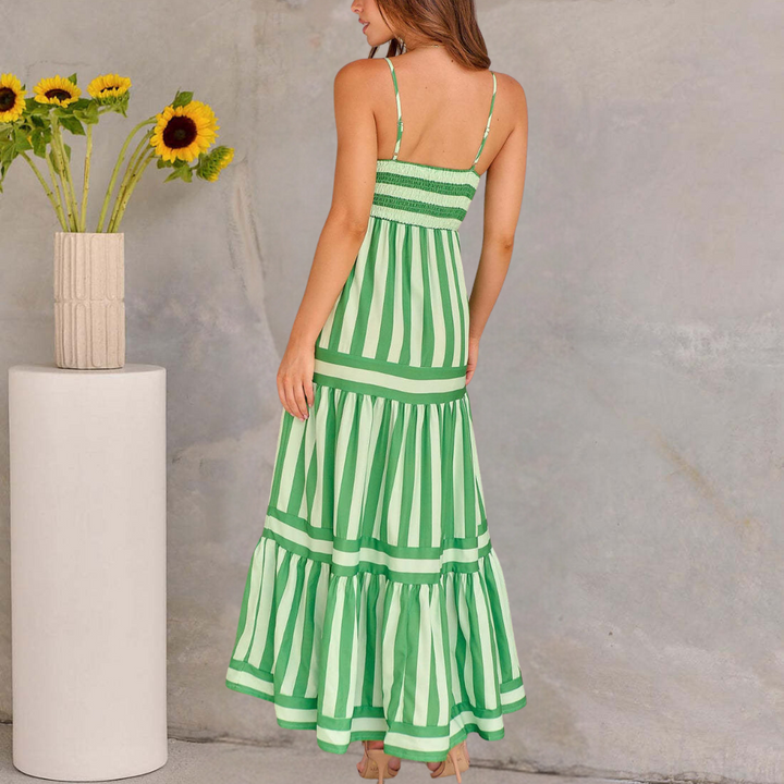 Vrouw in groene gestreepte zomerjurk met spaghettibandjes, staand naast een vaas met zonnebloemen op een betonnen achtergrond.