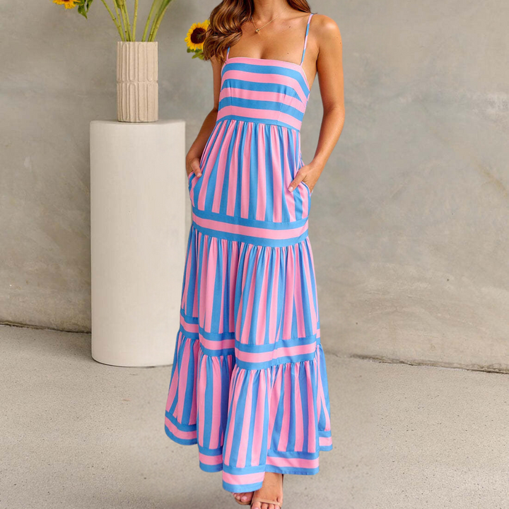 Vrouw in roze-blauwe gestreepte zomerjurk met zakken, staand voor een vaas met zonnebloemen. Perfecte outfit voor zomerse dagen.