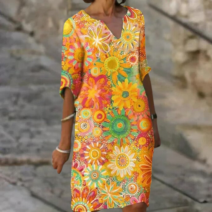 Vrouw in kleurrijke bloemenjurk met korte mouwen, oranje en gele bloemenprint, zomermode, casual stijl, buitenomgeving.
