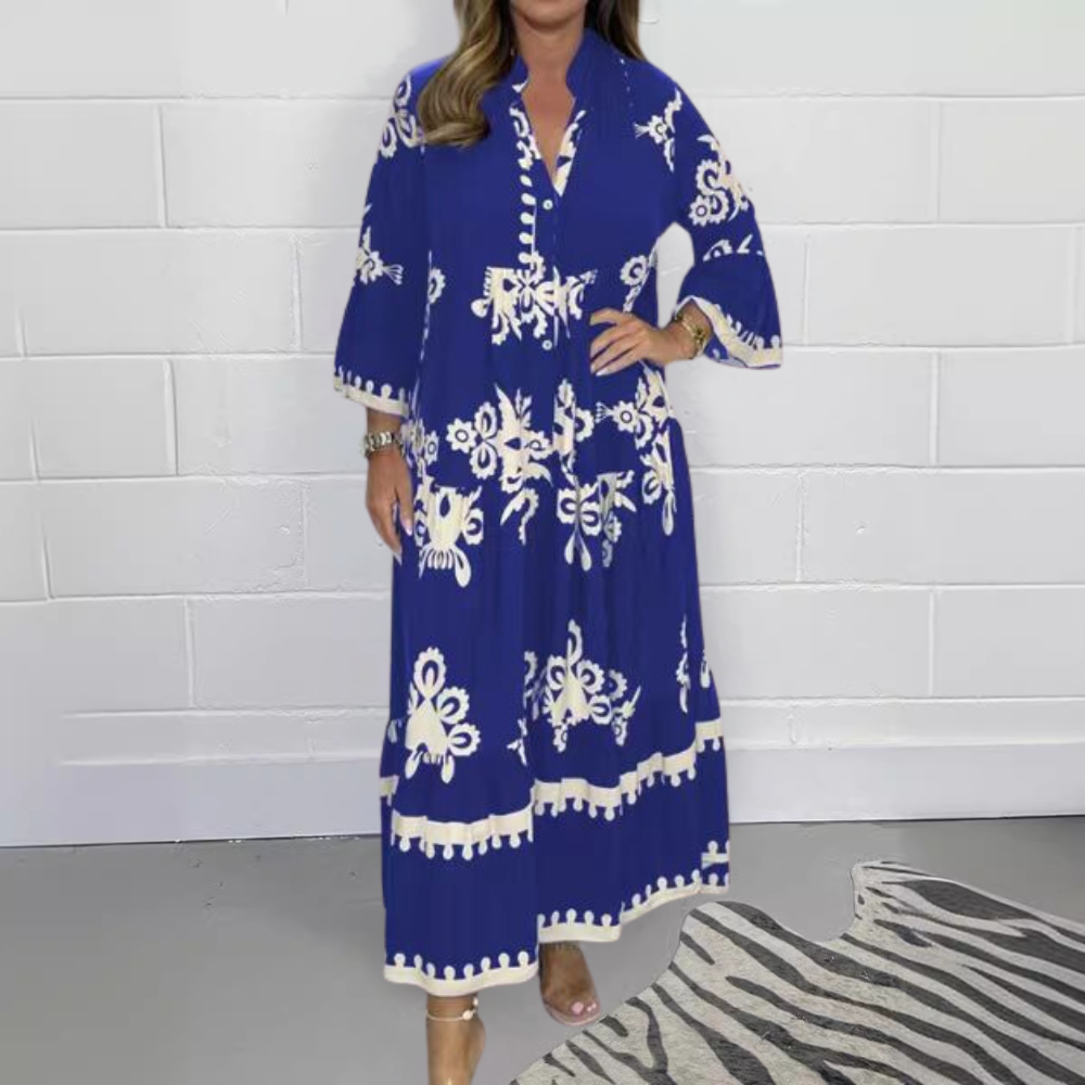 Vrouw in blauwe maxi-jurk met witte bloemenprint, driekwart mouwen en knoopsluiting, tegen witte bakstenen muur. Modieuze dameskleding.