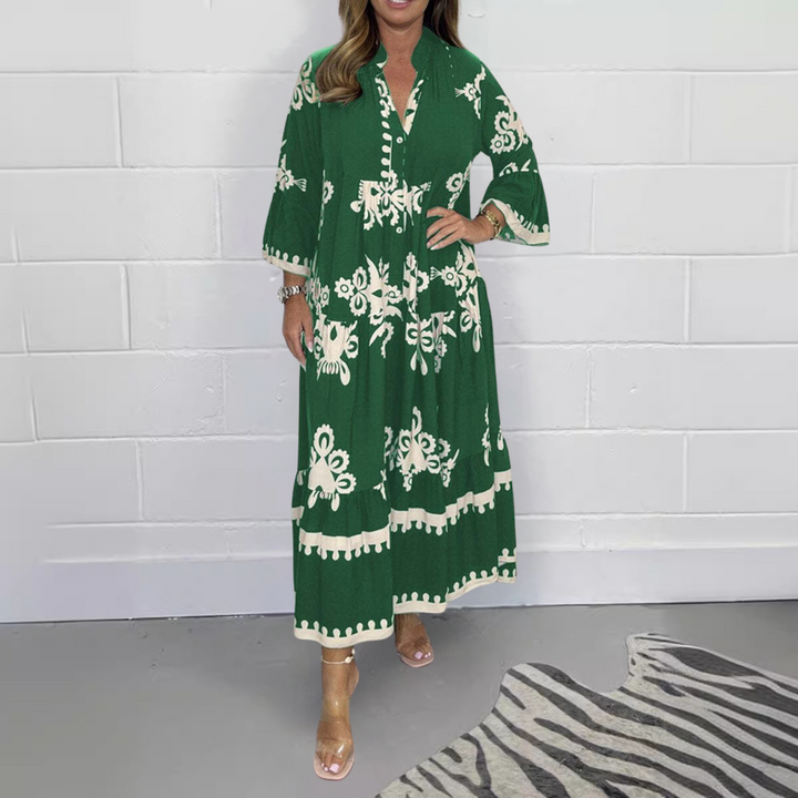 Vrouw in donkergroene maxi-jurk met witte bloemenprint, staand voor witte muur. Trendy damesmode, bohemian stijl, comfortabele pasvorm.