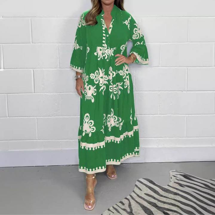Vrouw in groene boho-jurk met witte bloemenprint, staand voor witte bakstenen muur. Trendy zomeroutfit, casual en elegant.