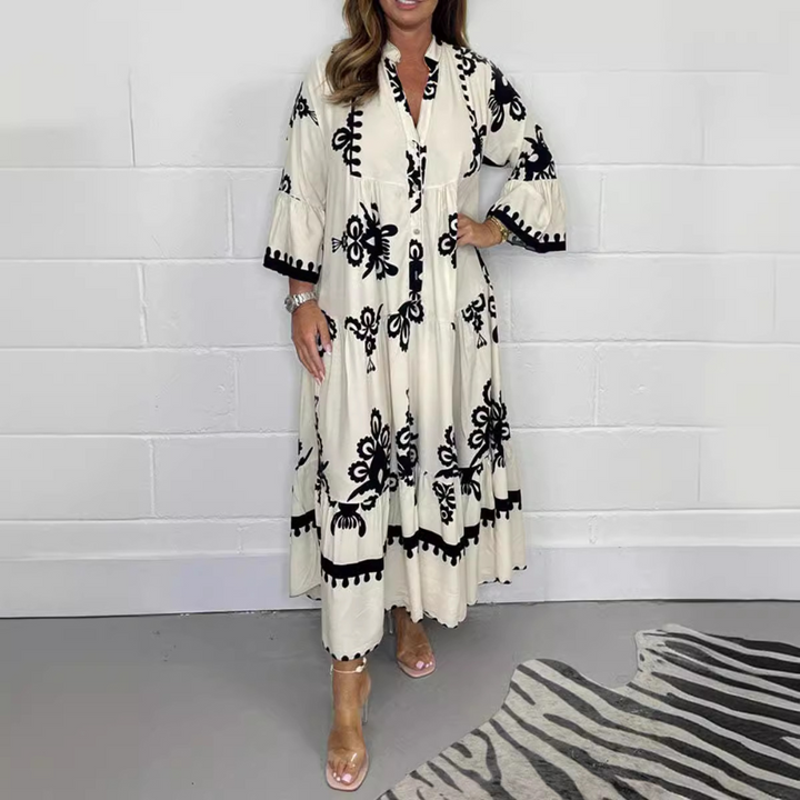 Vrouw in witte maxi-jurk met zwarte bloemenprint, staand op grijze vloer met zebraprint tapijt. Trendy damesmode, bohemian stijl.