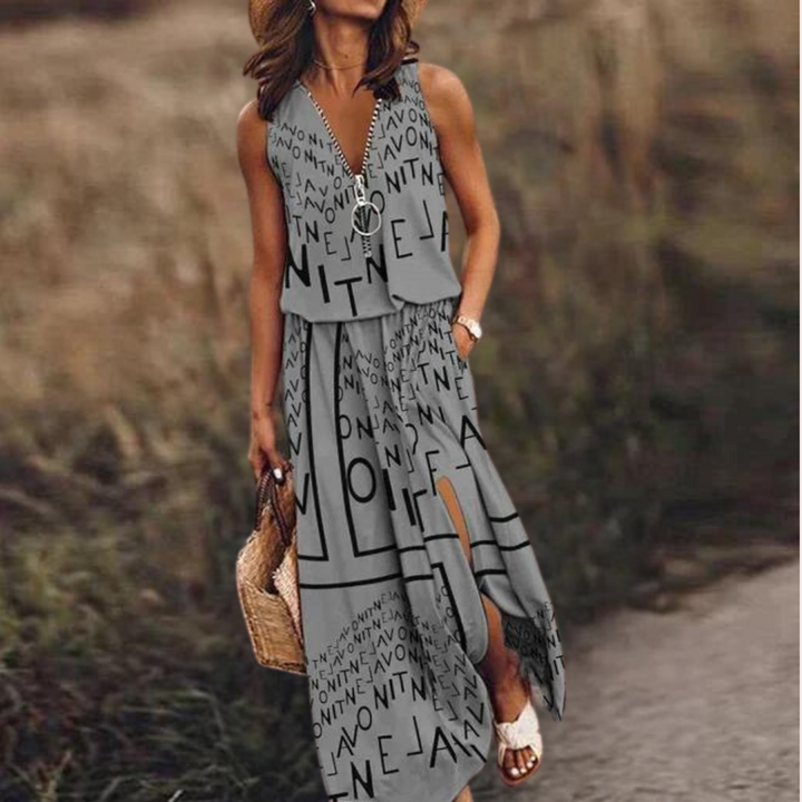 Vrouw in grijze zomerjurk met zwarte letters, draagt een strooien tas en witte sandalen, staat buiten op een grasveld. Trendy damesmode, casual stijl.