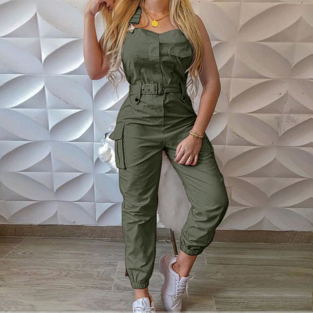 Vrouw in legergroene jumpsuit met riem, zakken en witte sneakers, tegen een moderne geometrische muur. Modieuze dameskleding, casual stijl.