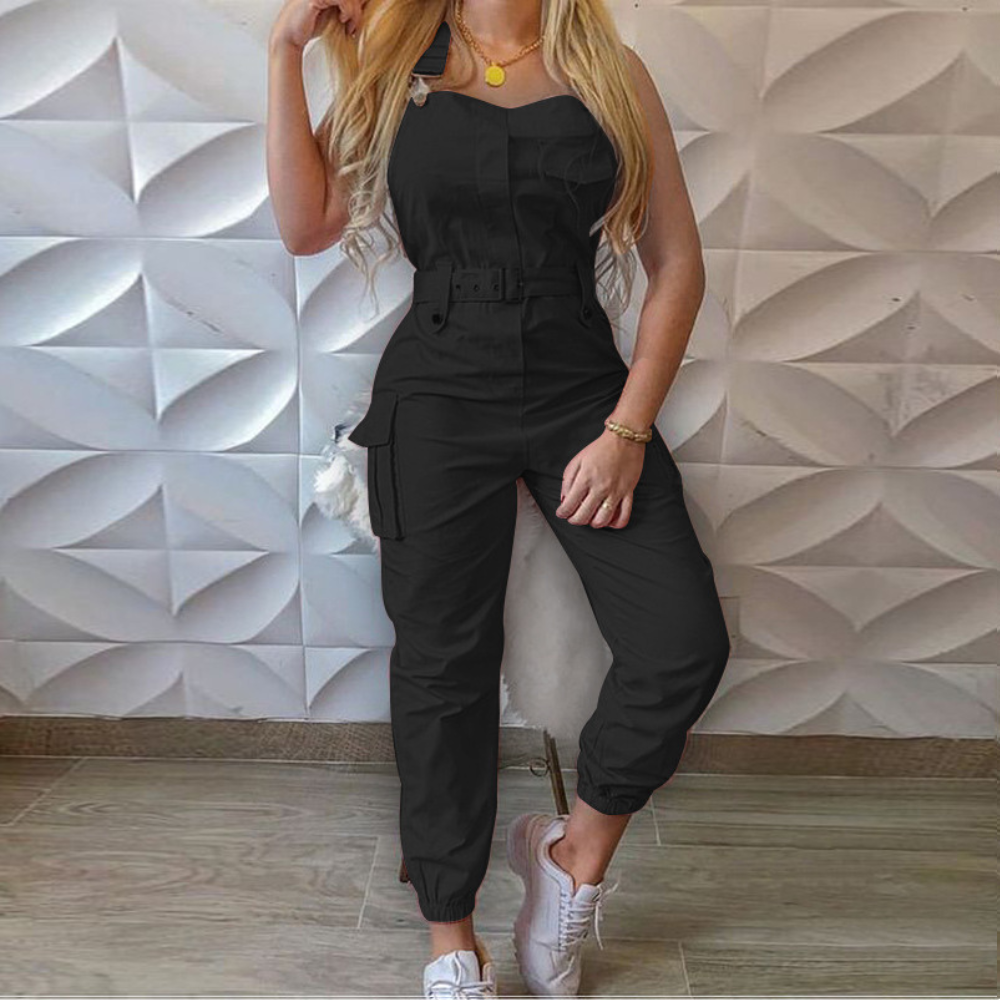 Vrouw in zwarte jumpsuit met riem, lange blonde haren, witte sneakers, tegen geometrische witte muur. Modieuze dameskleding, casual stijl.