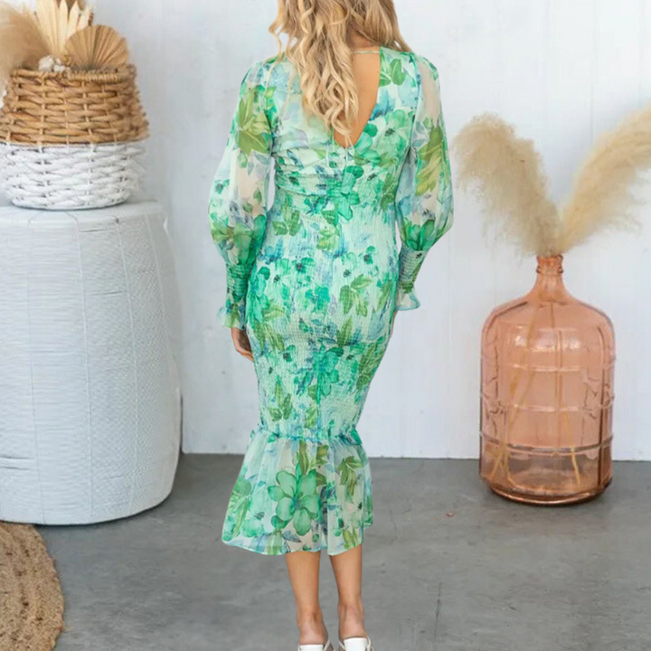 Vrouw in groene bloemenjurk met lange mouwen, staand in een moderne kamer met decoratieve mand en vaas. Elegante mode, lente/zomer stijl.