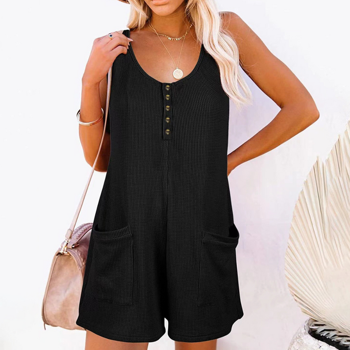 Vrouw in zwarte, mouwloze playsuit met knoopsluiting en zakken, draagt een beige hoed en tas. Zomermode, casual outfit, strandkleding.