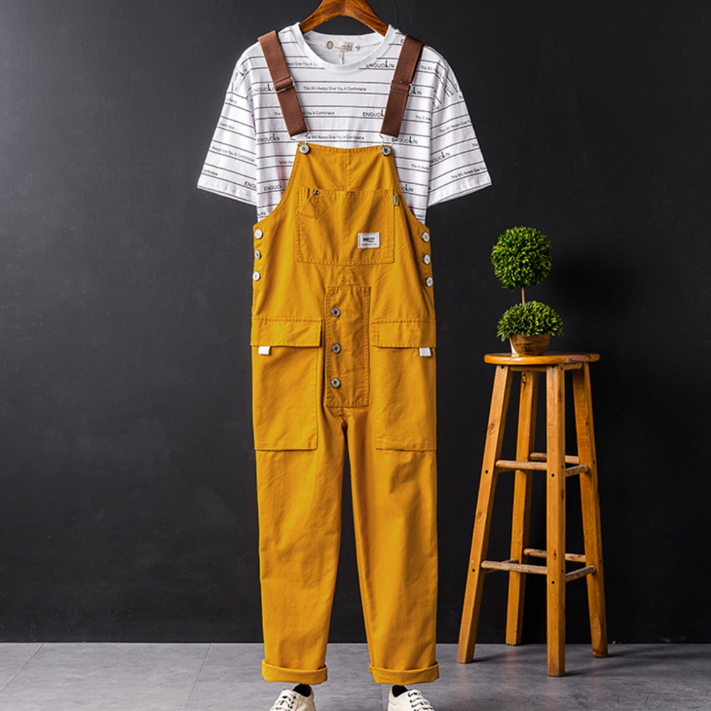Mosterdgele tuinbroek met witte gestreepte T-shirt, hangend aan een houten kruk, tegen een zwarte achtergrond. Modieuze casual outfit.