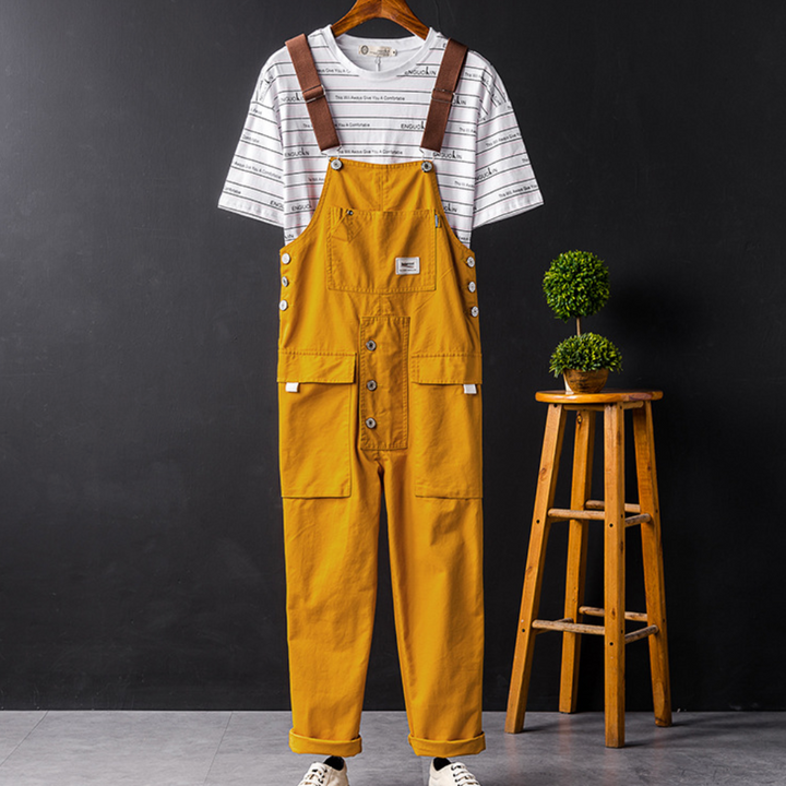 Mosterdgele tuinbroek met witte gestreepte T-shirt, hangend aan een houten kruk, tegen een zwarte achtergrond. Modieuze casual outfit.