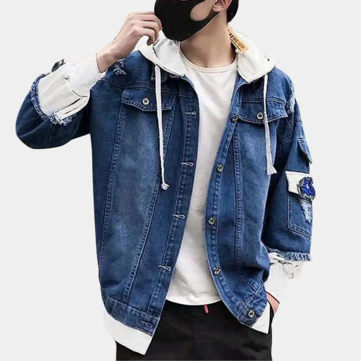 Man in donkerblauw denim jack met capuchon, witte voering en patches, draagt een zwart mondkapje. Modieuze streetwear outfit.