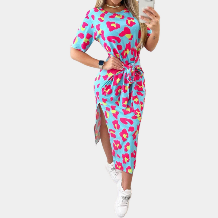 Vrouw in blauwe jurk met kleurrijke luipaardprint, korte mouwen en ceintuur, poseert met smartphone. Trendy damesmode, casual stijl.