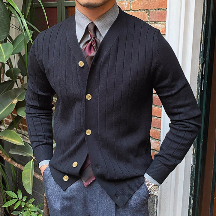 Man in zwarte cardigan met gouden knopen, grijze broek en stropdas, voor bakstenen muur. Stijlvolle herenkleding, formele outfit, mode.