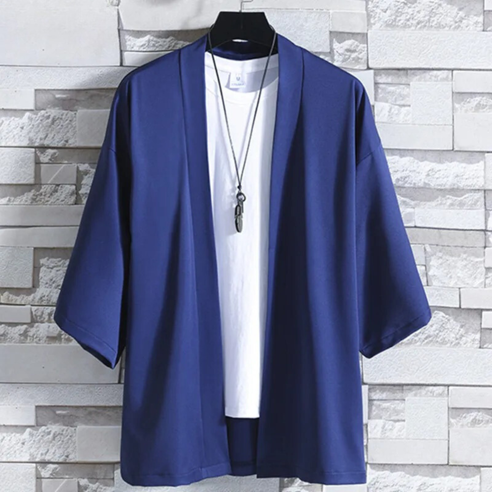 Blauwe kimono jas voor heren, casual stijl, driekwart mouwen, over een wit T-shirt, hangend aan een hanger tegen een stenen muur.