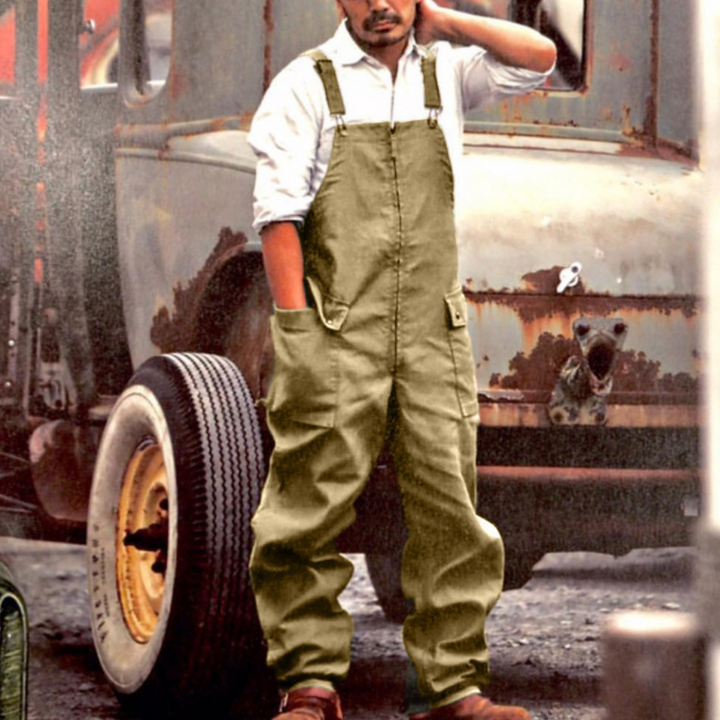 Man in legergroene tuinbroek voor vintage vrachtwagen, casual werkoutfit, stevige stof, praktische zakken, industriële achtergrond.