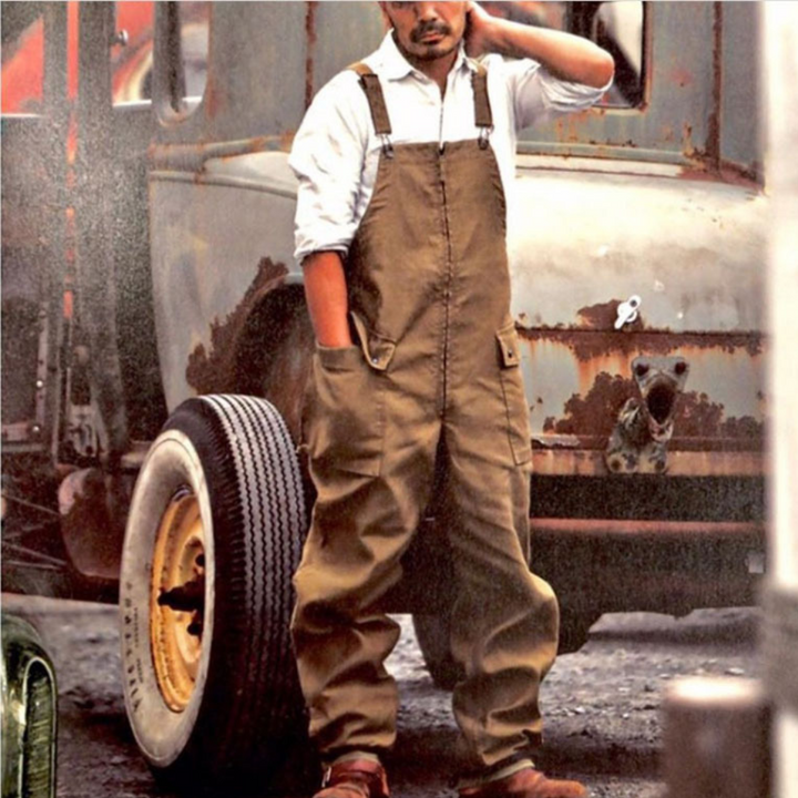 Man in bruine overall voor roestige vrachtwagen, werkoutfit, industriële stijl, vintage look, buitenomgeving, werklaarzen, casual kleding.