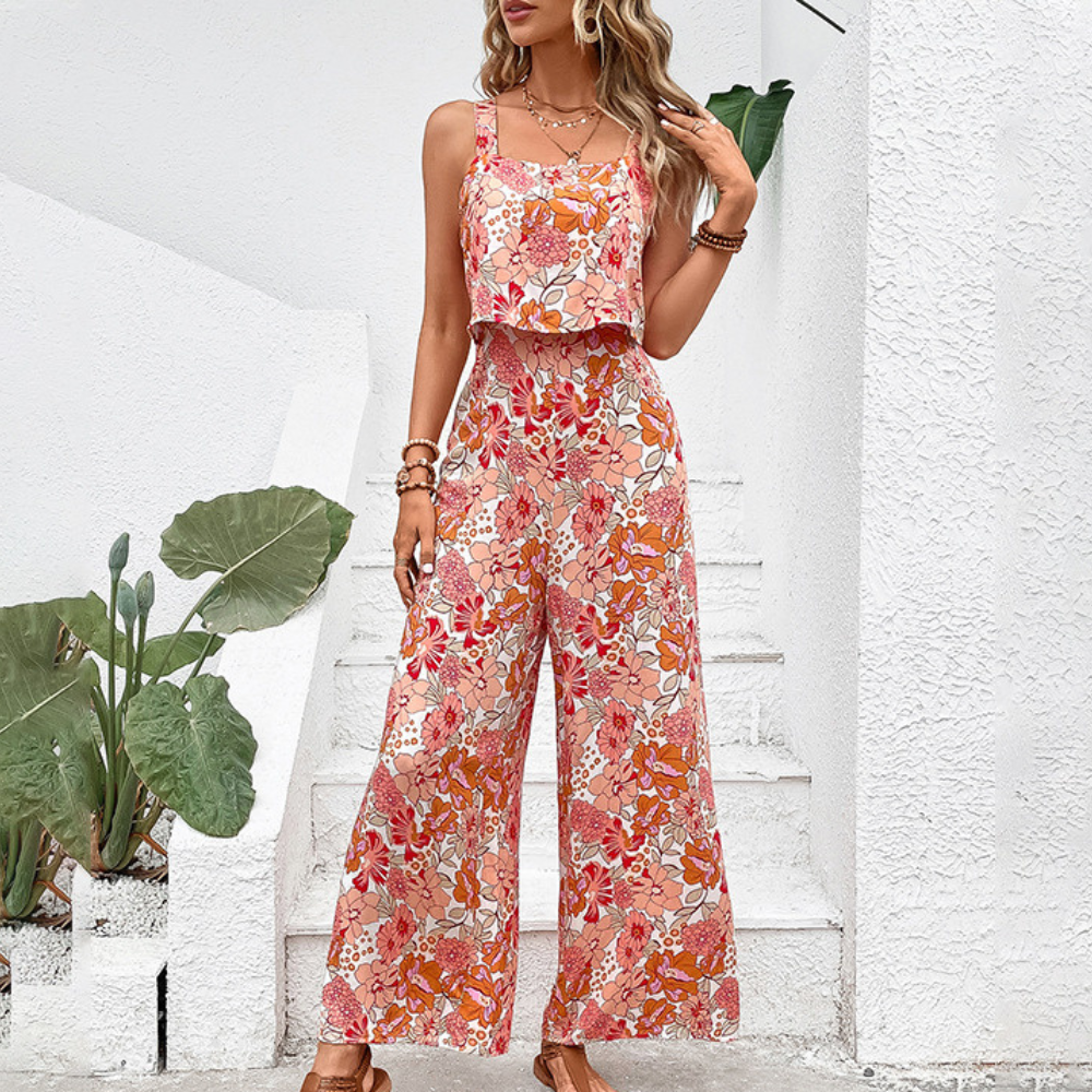 Vrouw in kleurrijke bloemen jumpsuit, oranje en roze tinten, zomermode, buiten bij witte muur met groene planten, trendy en stijlvol.