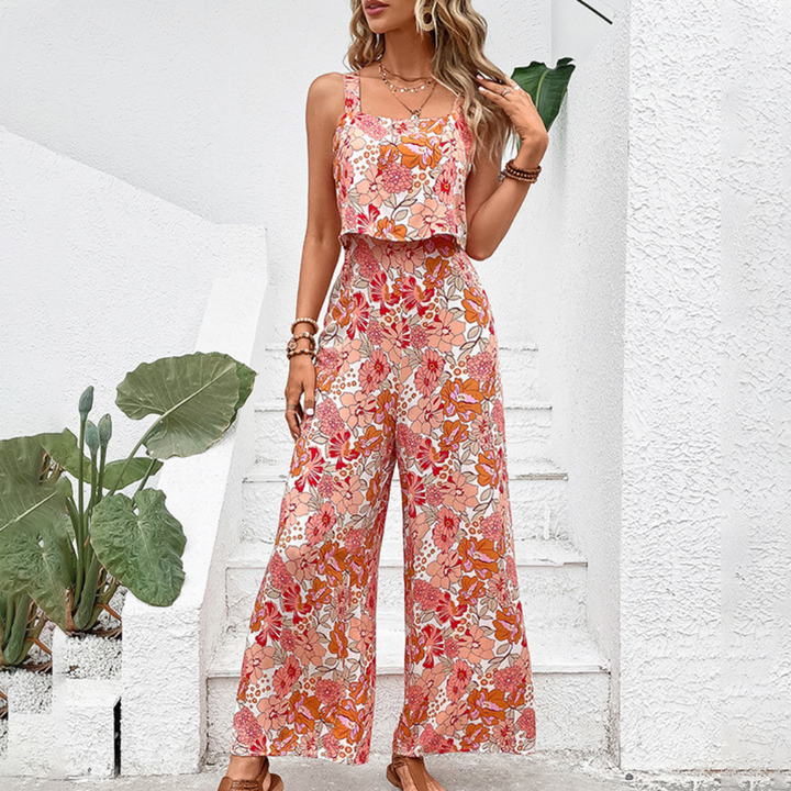 Vrouw in kleurrijke bloemen jumpsuit, oranje en roze tinten, zomermode, buiten bij witte muur met groene planten, trendy en stijlvol.