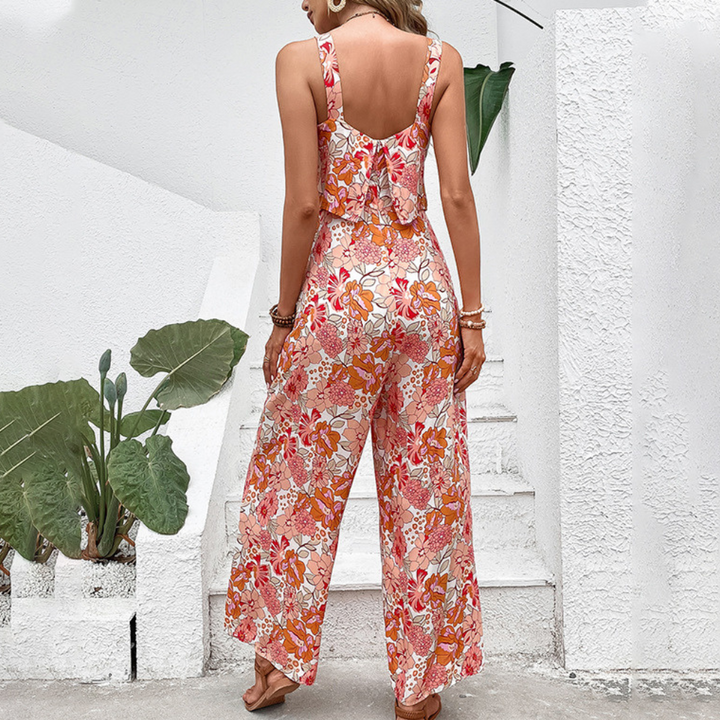 Vrouw in kleurrijke, bloemenprint jumpsuit met oranje en roze tinten, staand voor witte muur met groene planten. Zomermode, casual outfit.