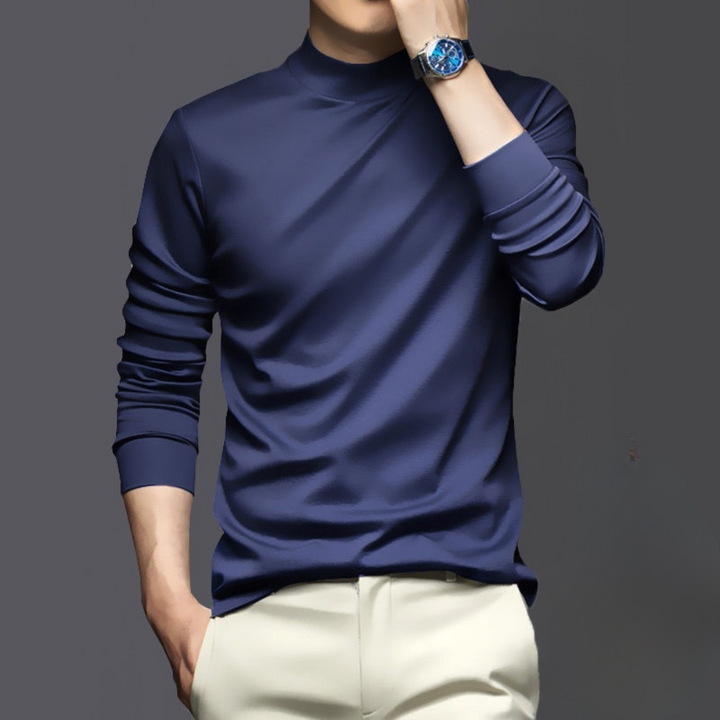 Man in donkerblauwe coltrui met opgerolde mouwen, draagt een luxe blauw horloge, poseert tegen een grijze achtergrond. Stijlvolle herenkleding.