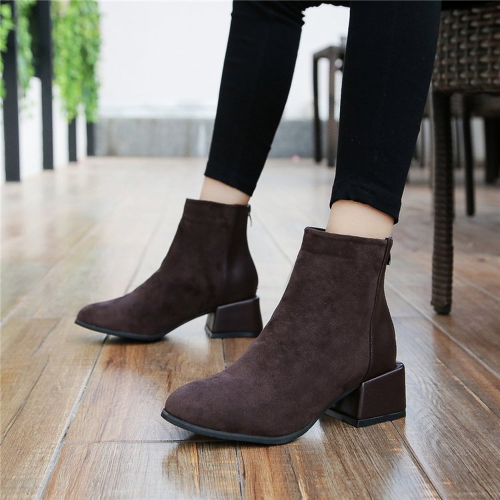 Bruine suède enkellaarsjes met blokhak, gedragen door model op houten vloer. Trendy damesmode, comfortabele herfst- en winterlaarzen.