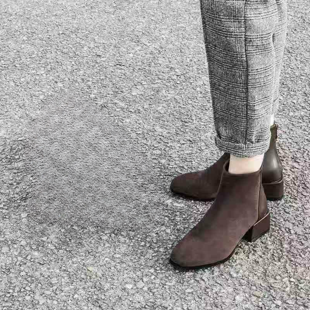 Donkerbruine suède enkellaarsjes met blokhak, gedragen met geruite broek op een grijze asfaltweg. Stijlvolle damesmode, herfst/winter schoenen.