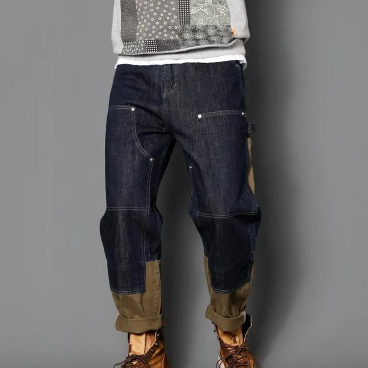 Donkerblauwe denim werkbroek met bruine kniestukken en opgerolde pijpen, gedragen door een persoon. Casual herenkleding, modieuze jeans.