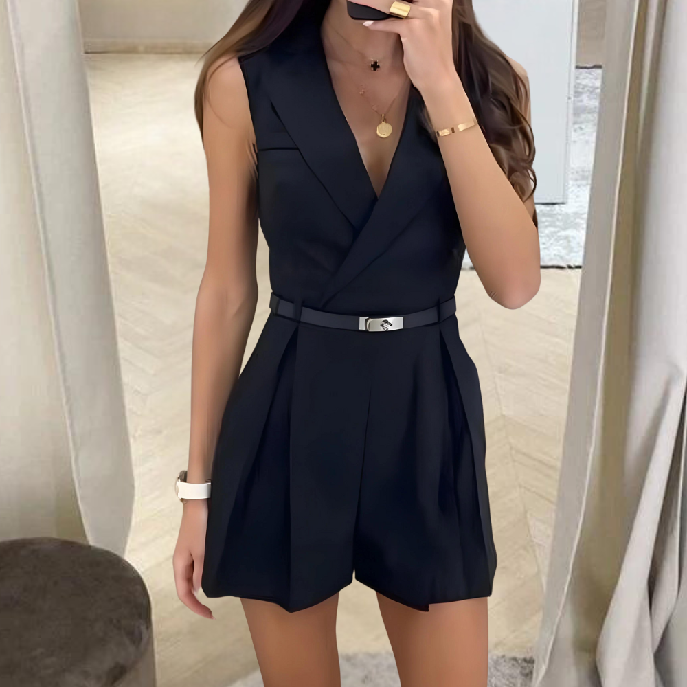Vrouw in elegante zwarte mouwloze jumpsuit met V-hals en riem, staand in moderne kamer. Perfect voor chique avondkleding en mode-inspiratie.